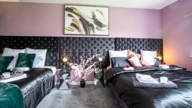 Ein modernes Schlafzimmer mit einem gepolsterten Kopfteil, zwei Betten mit dunkler Bettwäsche, einem großen abstrakten Gemälde und einer Vase mit Blumen.