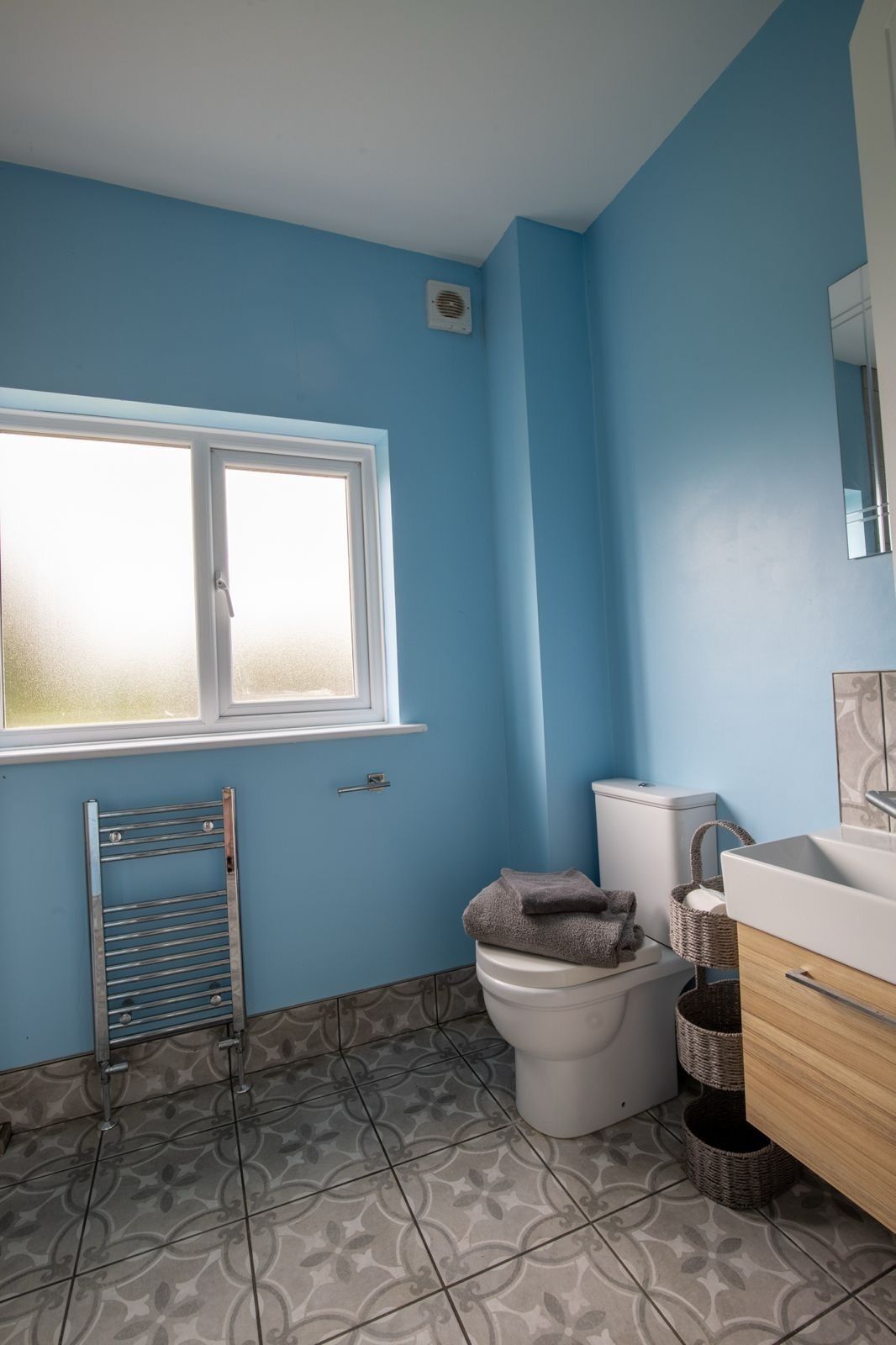 Et badeværelse med blå væg, et vindue, et toilet, et håndklædestativ og en vask med en kurv.