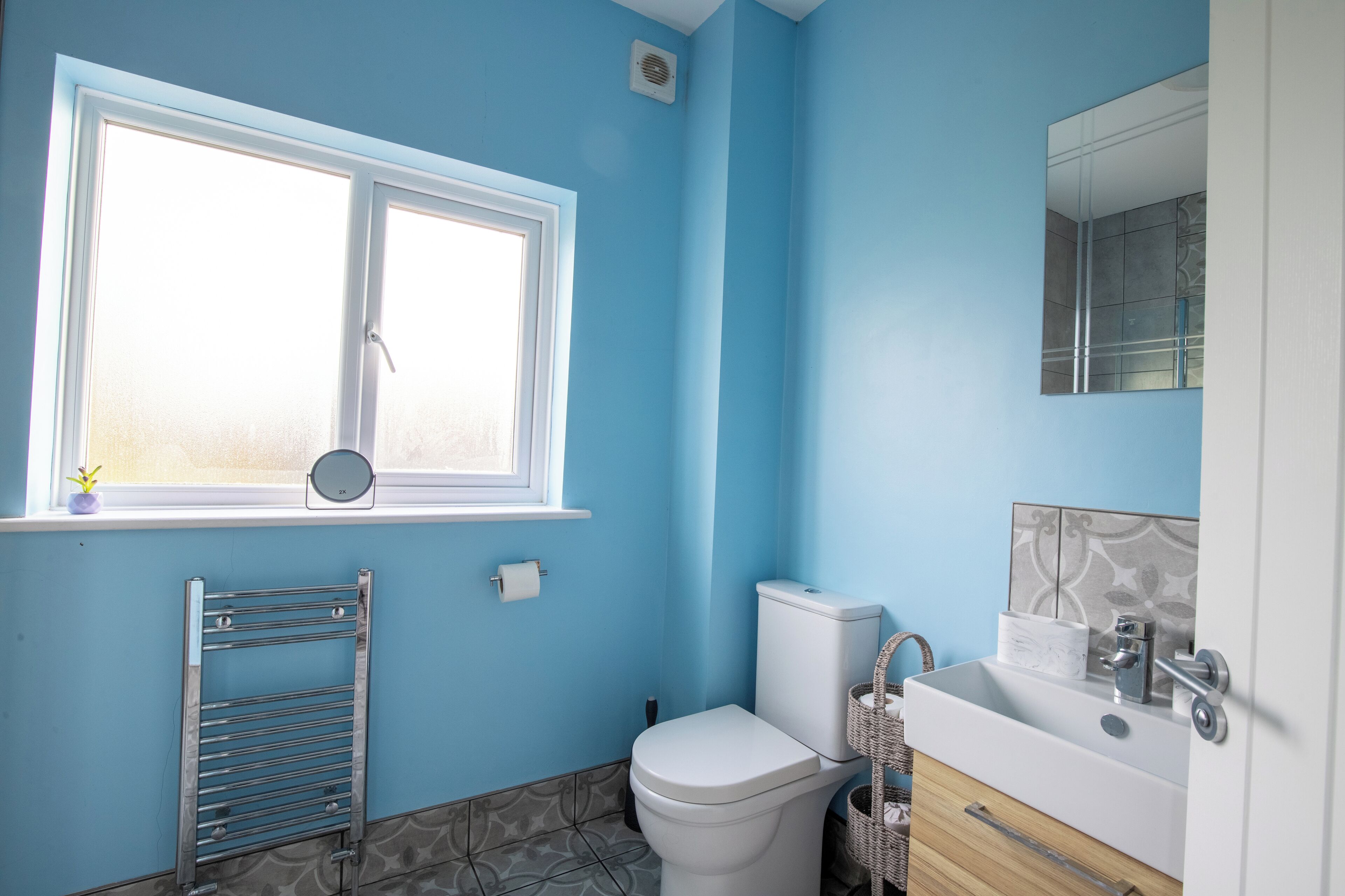 Une salle de bain avec des toilettes blanches, un lavabo avec un miroir et une fenêtre donnant sur l’extérieur.