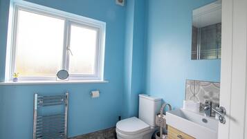 Une salle de bain avec des toilettes blanches, un lavabo avec un miroir et une fenêtre donnant sur l’extérieur.