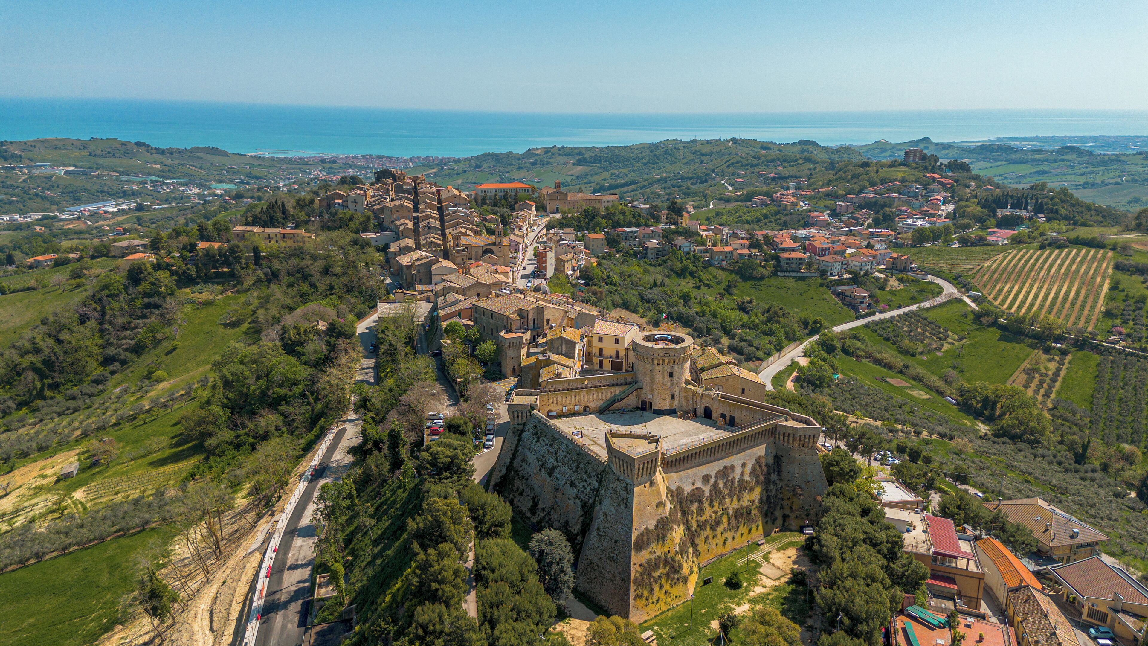 Vista aérea de un pueblo histórico con un castillo destacado, rodeado de exuberante vegetación y campos.