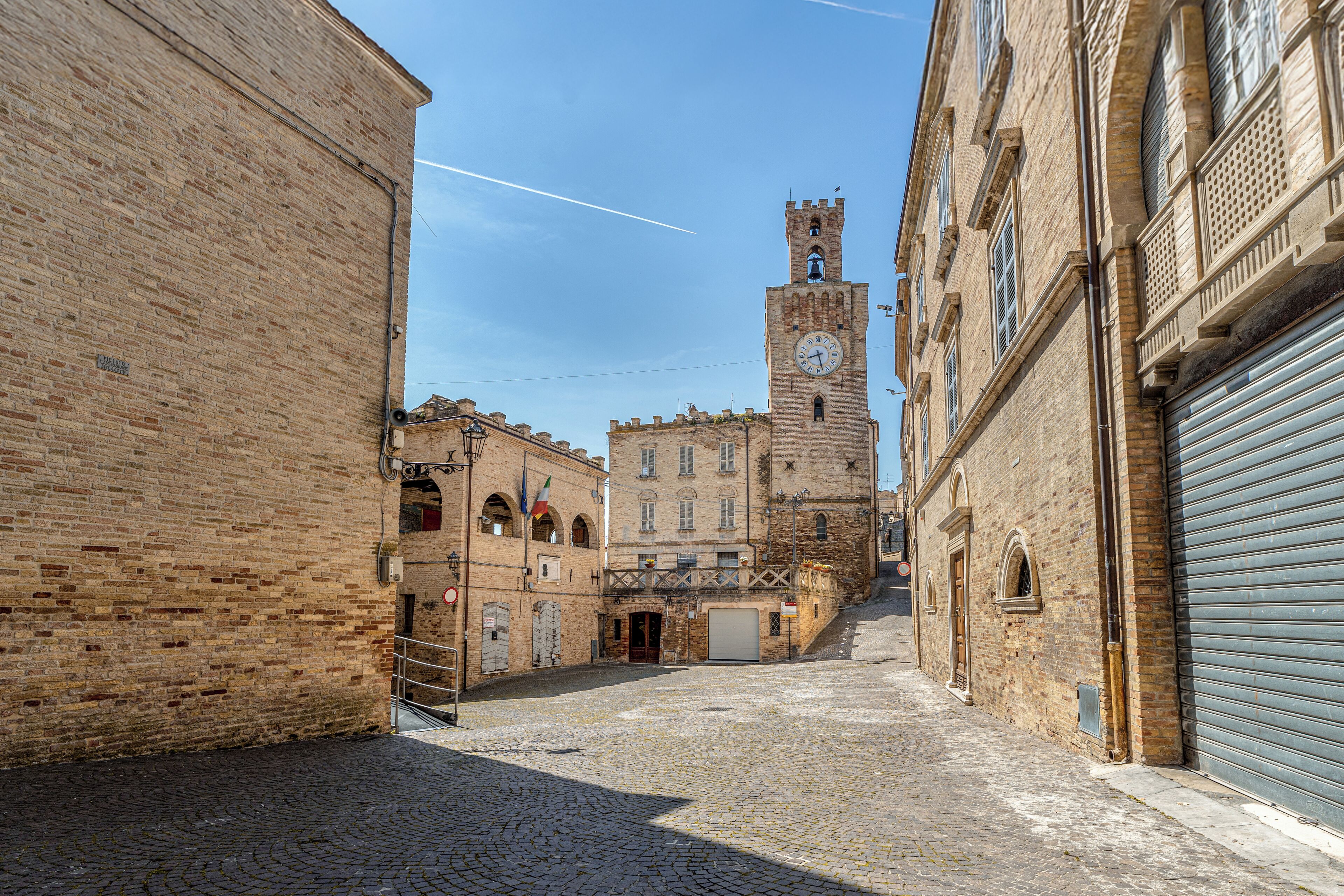 A cobblestone street leading to a historic building with a clock tower, arched doorways, and a closed shutter on the right.