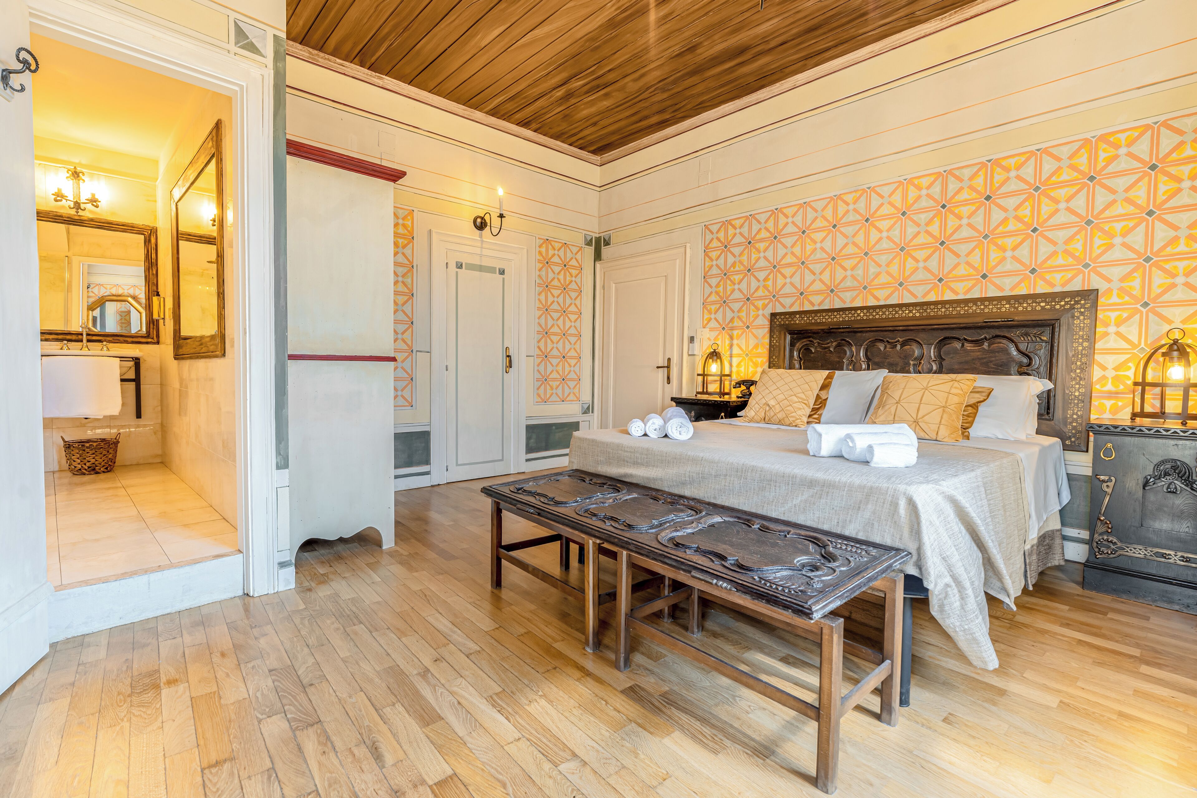 Un dormitorio con suelo de madera, una cama con almohadas, un banco y una mesita de noche con lámpara.