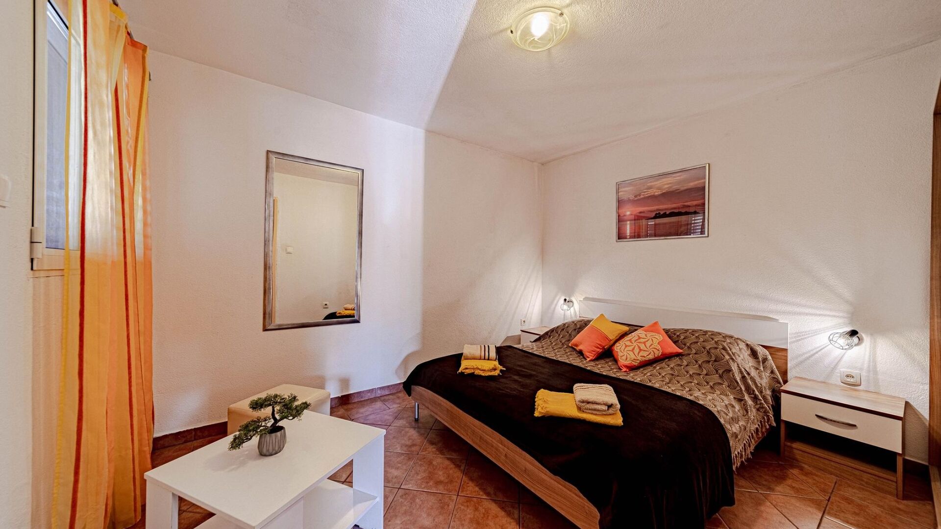 A bedroom with a bed, bedside tables, a desk, a chair, a potted plant, and a painting on the wall.