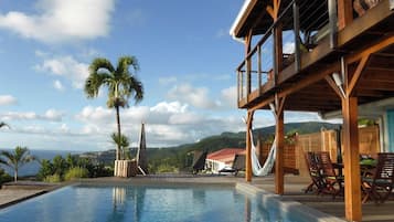Une piscine dotée d’une terrasse en bois et de chaises longues, offrant une vue sur un paysage montagneux parsemé de palmiers.