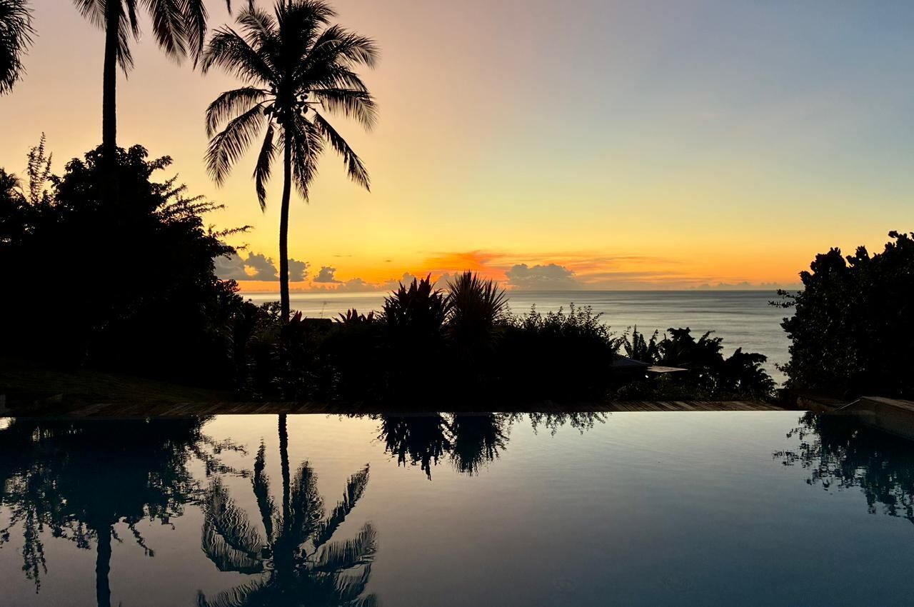 Une vue sur le coucher de soleil depuis une piscine à débordement, avec des palmiers et un ciel dégagé.