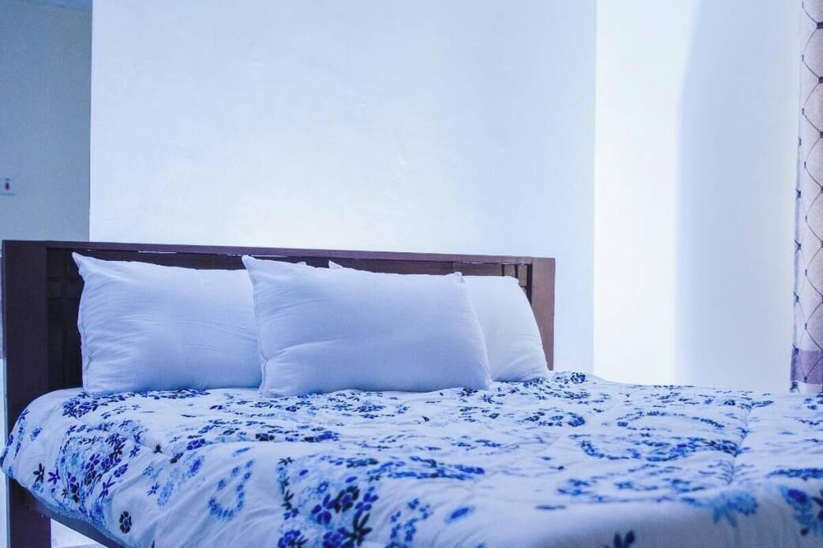 A bed with white pillows and a blue floral bedspread in a room with a wooden headboard and sheer curtains.