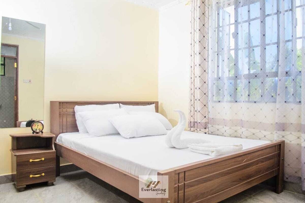 A neatly made bed with white linens and a swan-shaped towel, a wooden nightstand with a clock, and a window with patterned curtains.