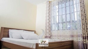 A neatly made bed with white linens, a wooden headboard, and a window with patterned curtains. - Everlasting Gardens Kitui (Kitui)