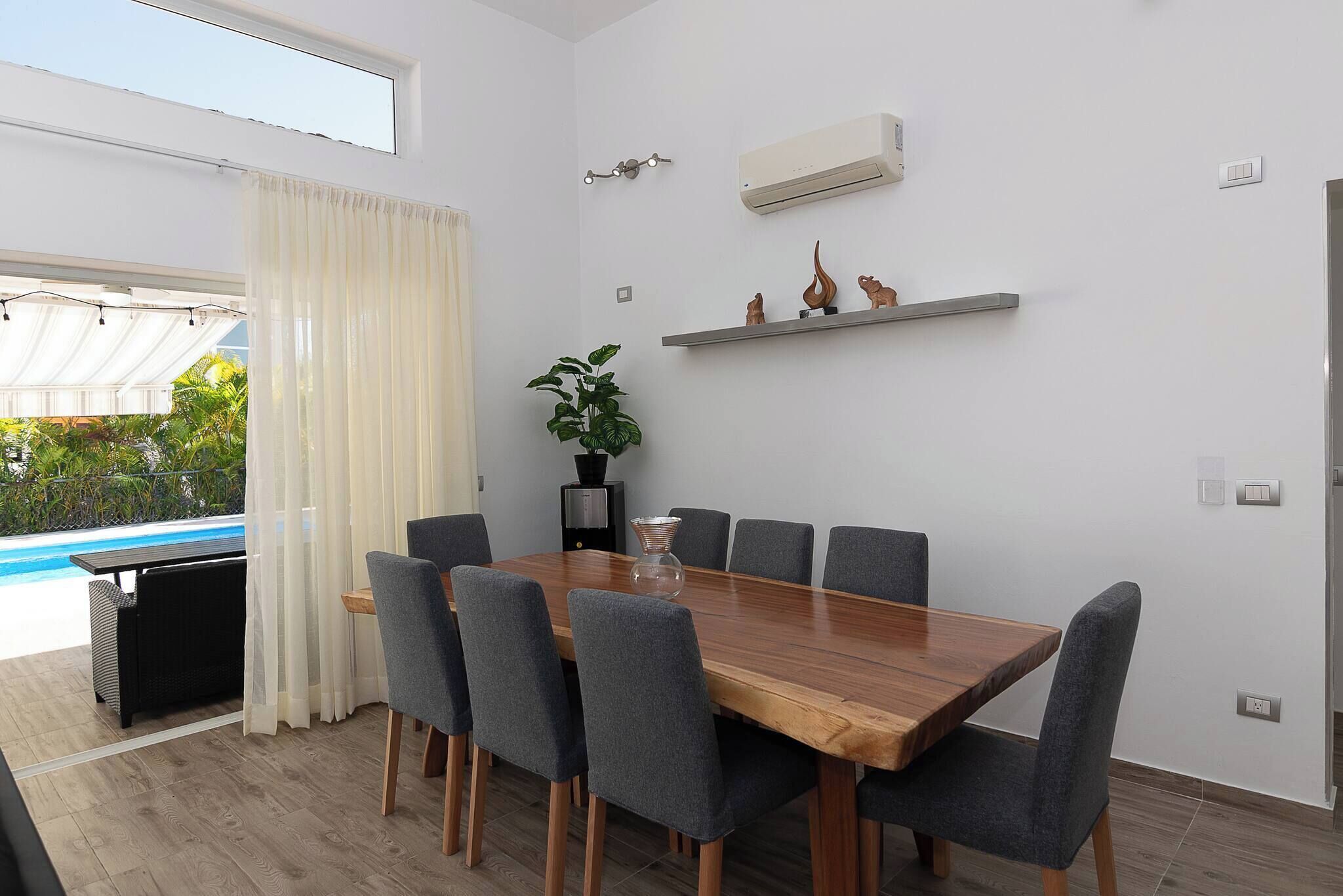 A modern dining room with a wooden table and grey chairs, a potted plant, and a pool visible through the window.