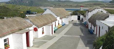 A row of white buildings with thatched roofs, a paved courtyard, and a view of hills and water in the distance.