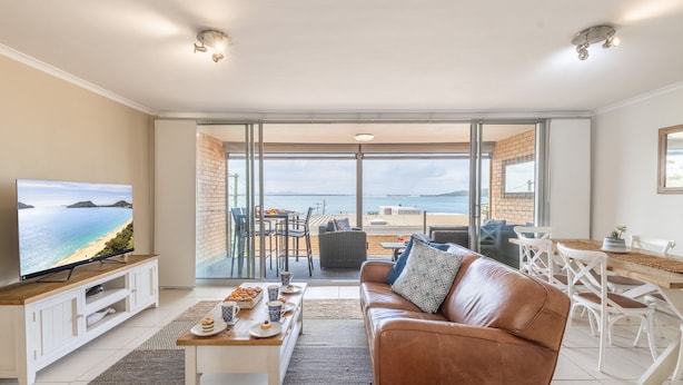 Ett vardagsrum med en tv, en soffa, ett matbord med stolar och utsikt över havet genom stora glasdörrar.