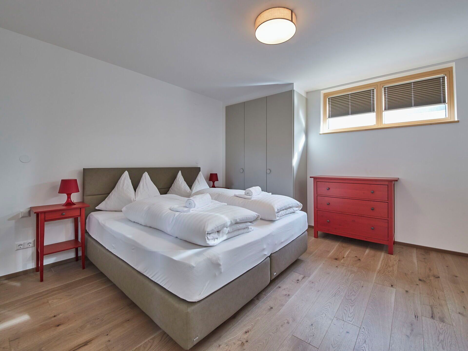 A bedroom with a bed, a red nightstand, a red dresser, and a window with blinds.