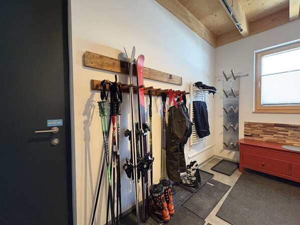 Une pièce servant de local à skis, où l’on peut ranger les skis et les chaussures de ski sur des supports, avec un banc en bois et une fenêtre équipée de stores.
