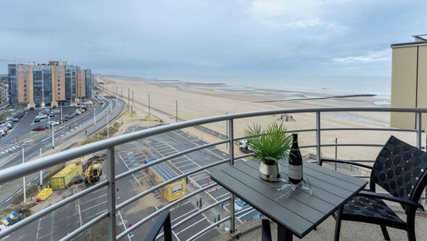A balcony with a table, chairs, and a bottle of wine. A view of a beach and a road with cars is visible. - Cosy apartment for 4 people with WIFI, TV, balcony and panoramic view (Bruges)