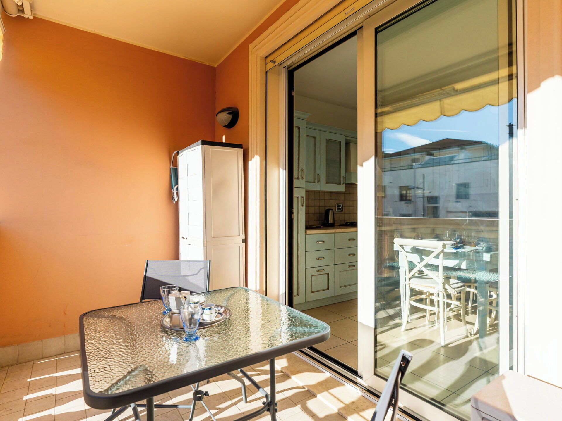 Een balkon met een glazen tafel en stoelen, een koelkast, en een keuken zichtbaar door de glazen deuren.