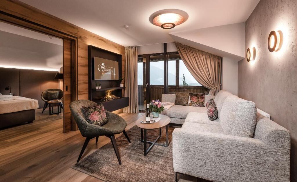 Une chambre d’hôtel moderne comprenant un lit, un canapé, un fauteuil, une petite table avec un vase et une grande fenêtre avec des rideaux.