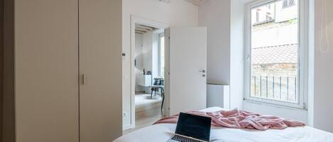Un dormitorio con cama, una computadora portátil, una ventana y una puerta que da a otra habitación.
