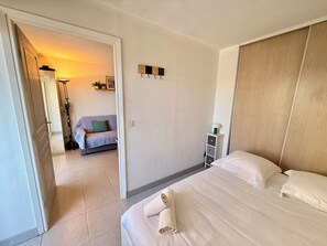 A hotel room with a bed, a small bedside table, a nightstand, a wall-mounted light, and a view of a hallway with a sofa and a lamp. - Apartment T2 Port Fréjus (Fréjus)