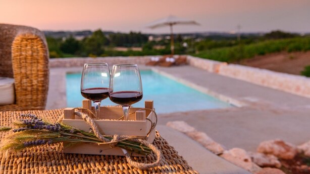 A poolside setting with two glasses of red wine on a woven mat, surrounded by a wicker chair and a wooden box. - Authentic Trulli Stay with Private Pool and Panoramic Views | Itria Valley (Martina Franca)