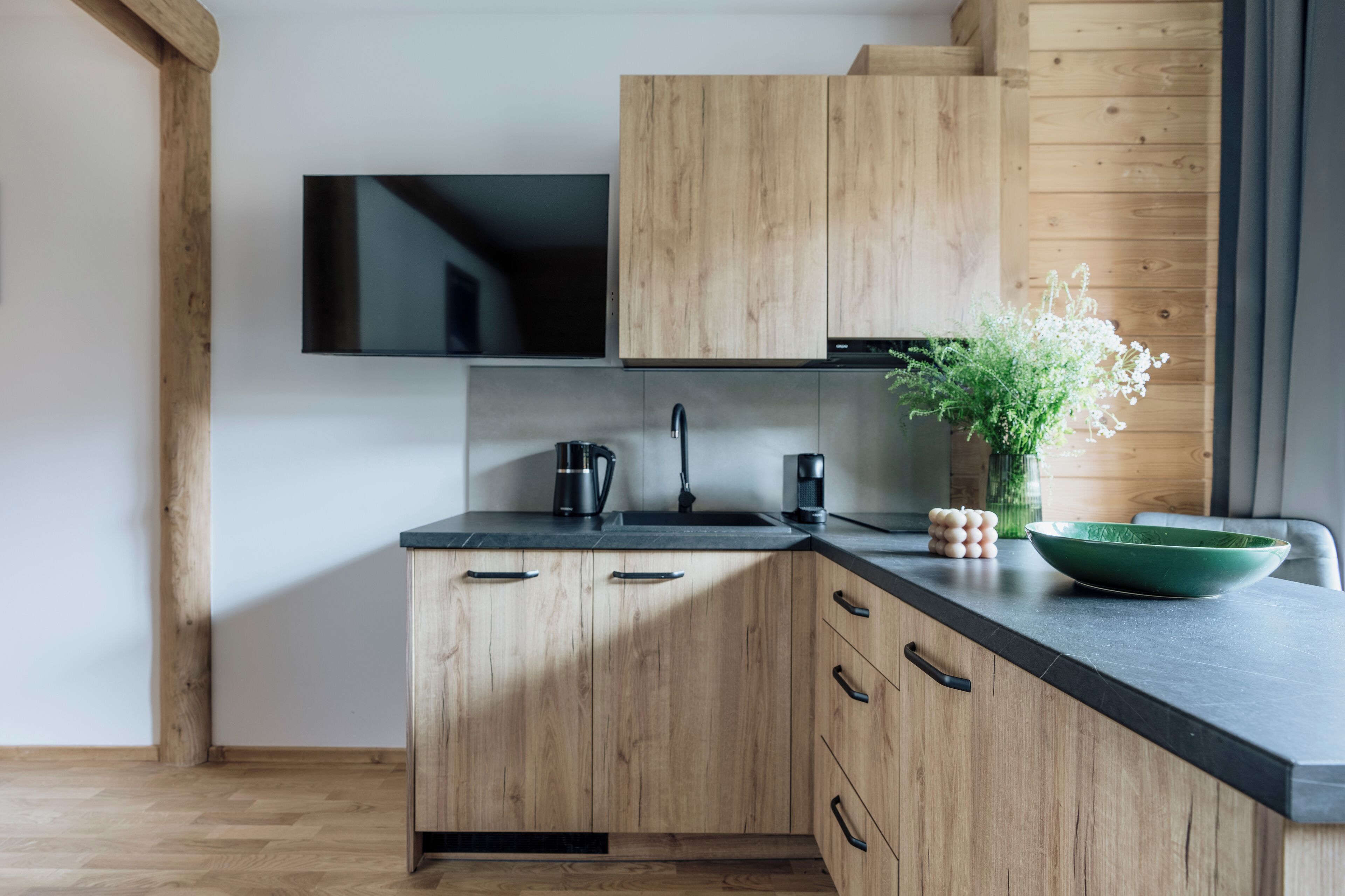 A modern kitchen with wooden cabinets, a black countertop, and a flat-screen TV mounted above the sink.