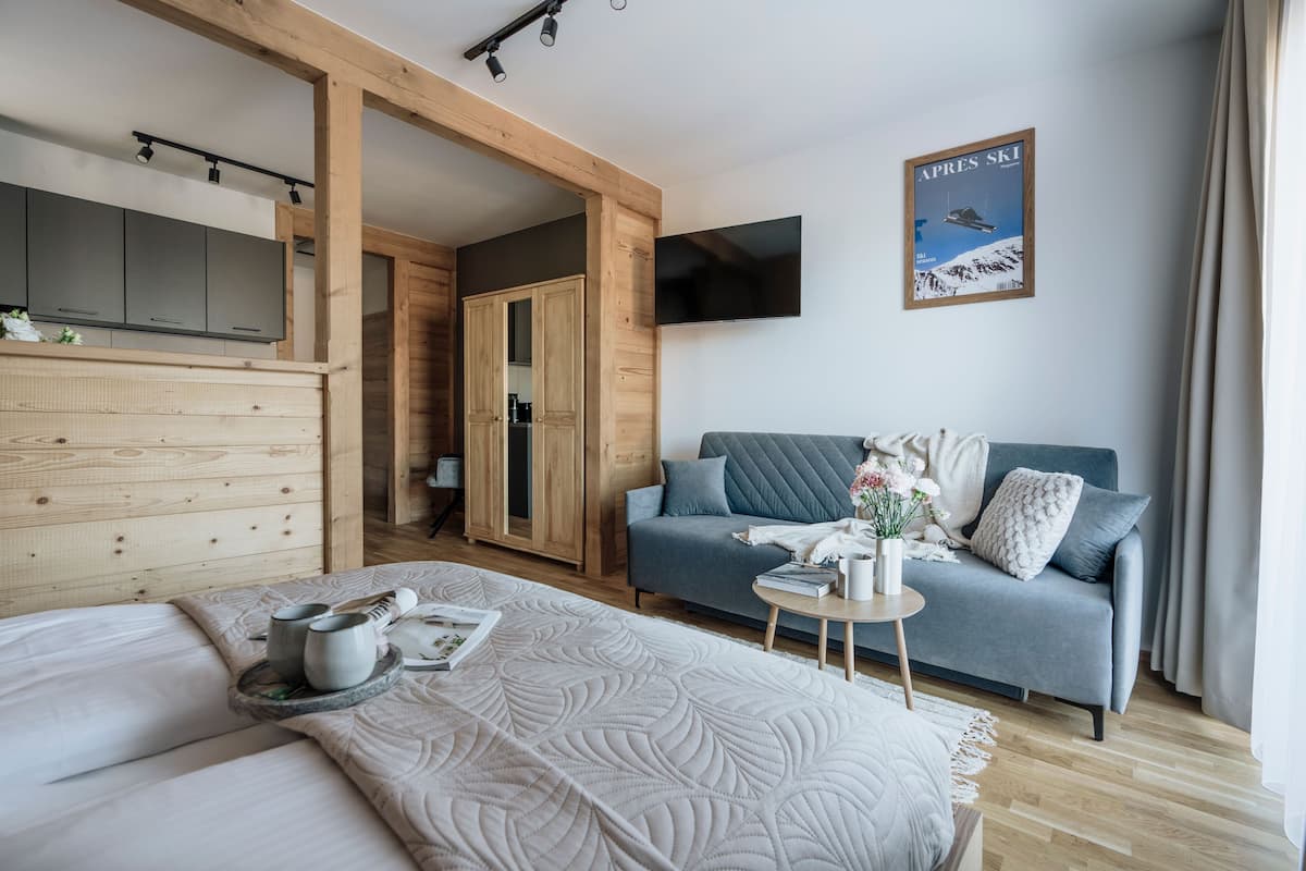 A modern hotel room with a wooden floor, a grey sofa, a bed with a white quilt, a small table with a vase, and a TV mounted on the wall.
