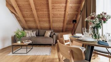 Um quarto acolhedor no estilo loft, com teto de madeira, sofá cinza, mesa de jantar posta para quatro pessoas e um vaso de flores.