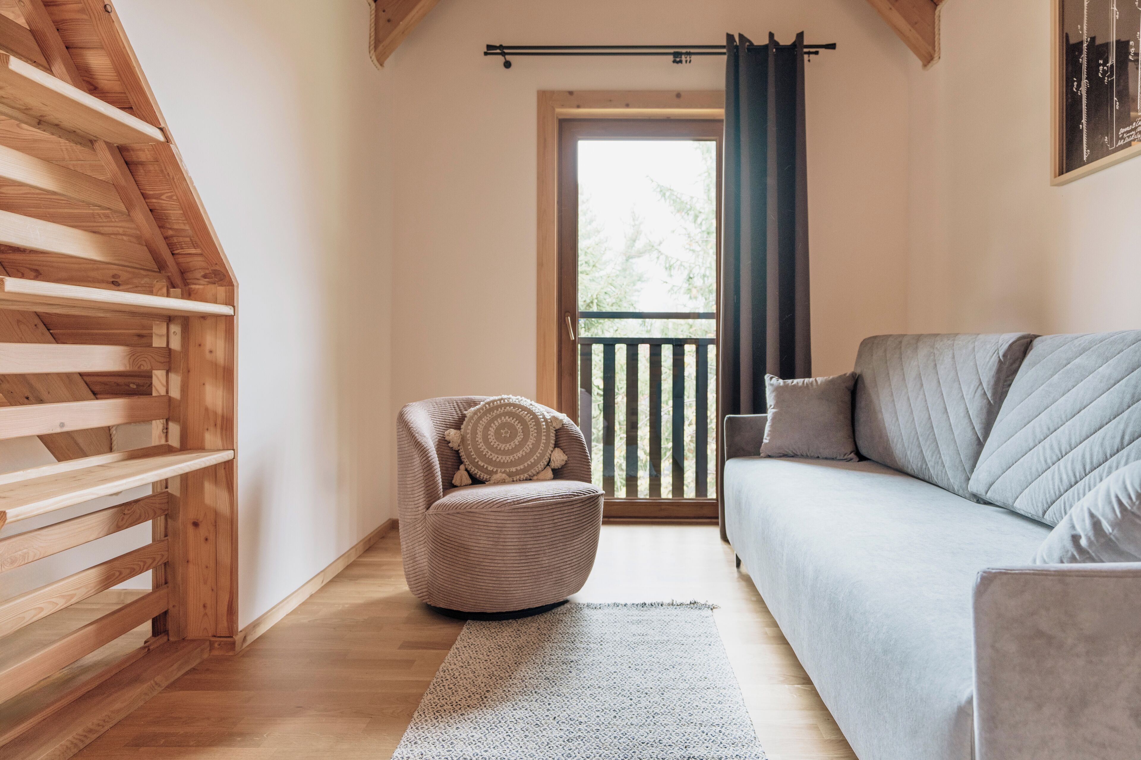 A cozy room with a wooden slatted ceiling, a grey sofa, a round chair with a cushion, and a view through a door to an outdoor balcony.