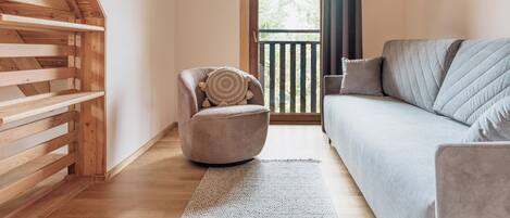 A cozy room with a wooden slatted ceiling, a grey sofa, a round chair with a cushion, and a view through a door to an outdoor balcony.