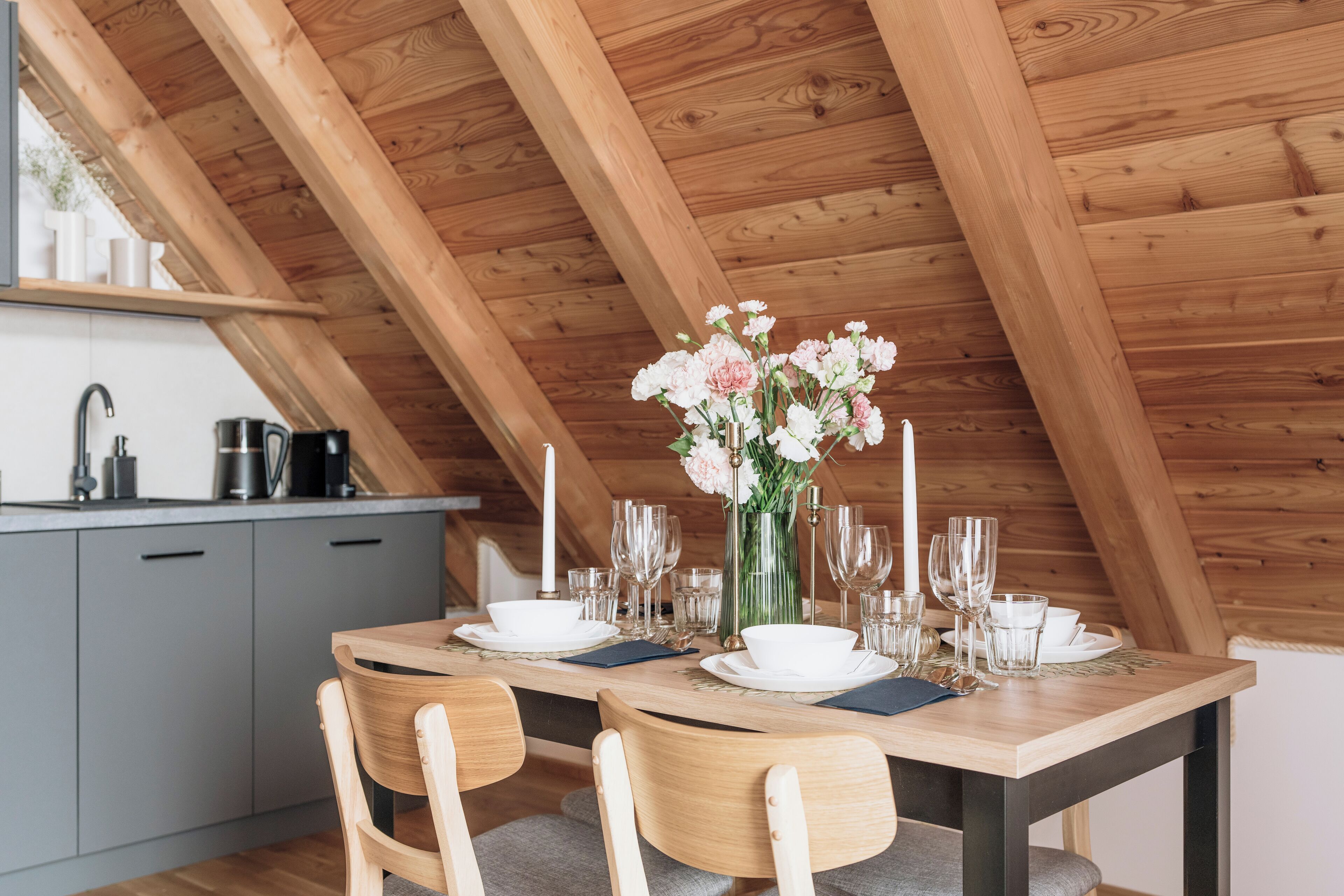 A dining area with a wooden table set for two, a vase of flowers, and a kitchen in the background.