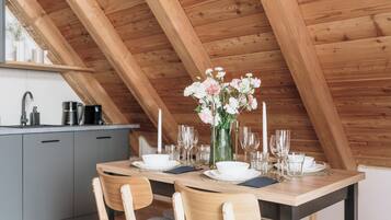 A dining area with a wooden table set for two, a vase of flowers, and a kitchen in the background.