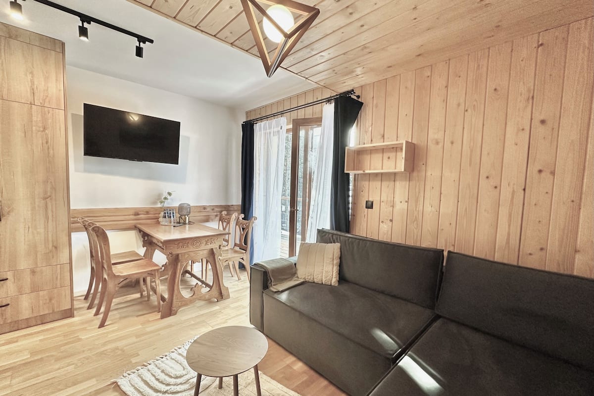 A cozy room with a wooden floor, a sofa, a dining table with chairs, and a TV mounted on the wall.