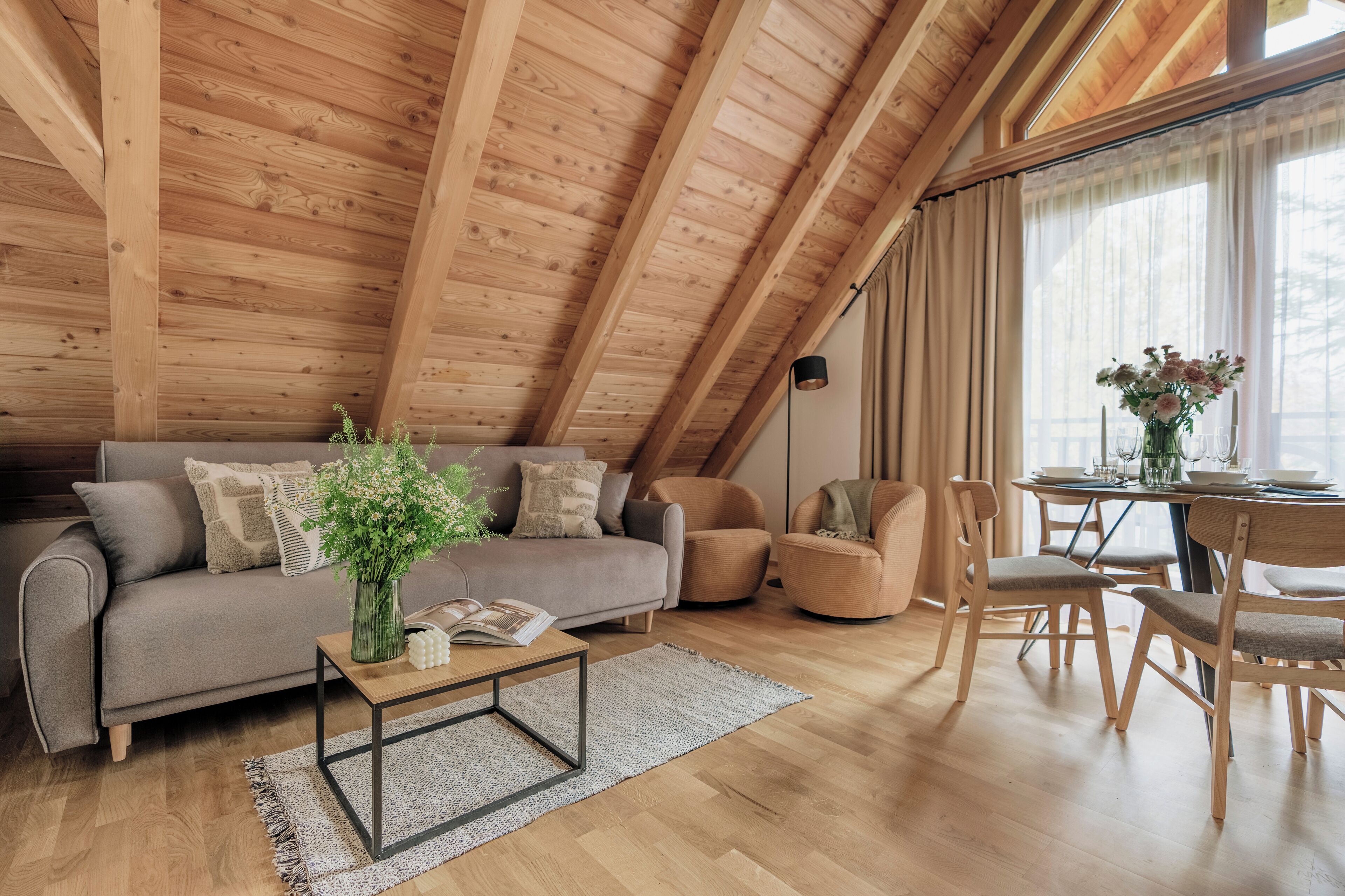 A cozy loft-style room with a wooden ceiling, a grey sofa, a coffee table, and a dining area with a table and chairs.
