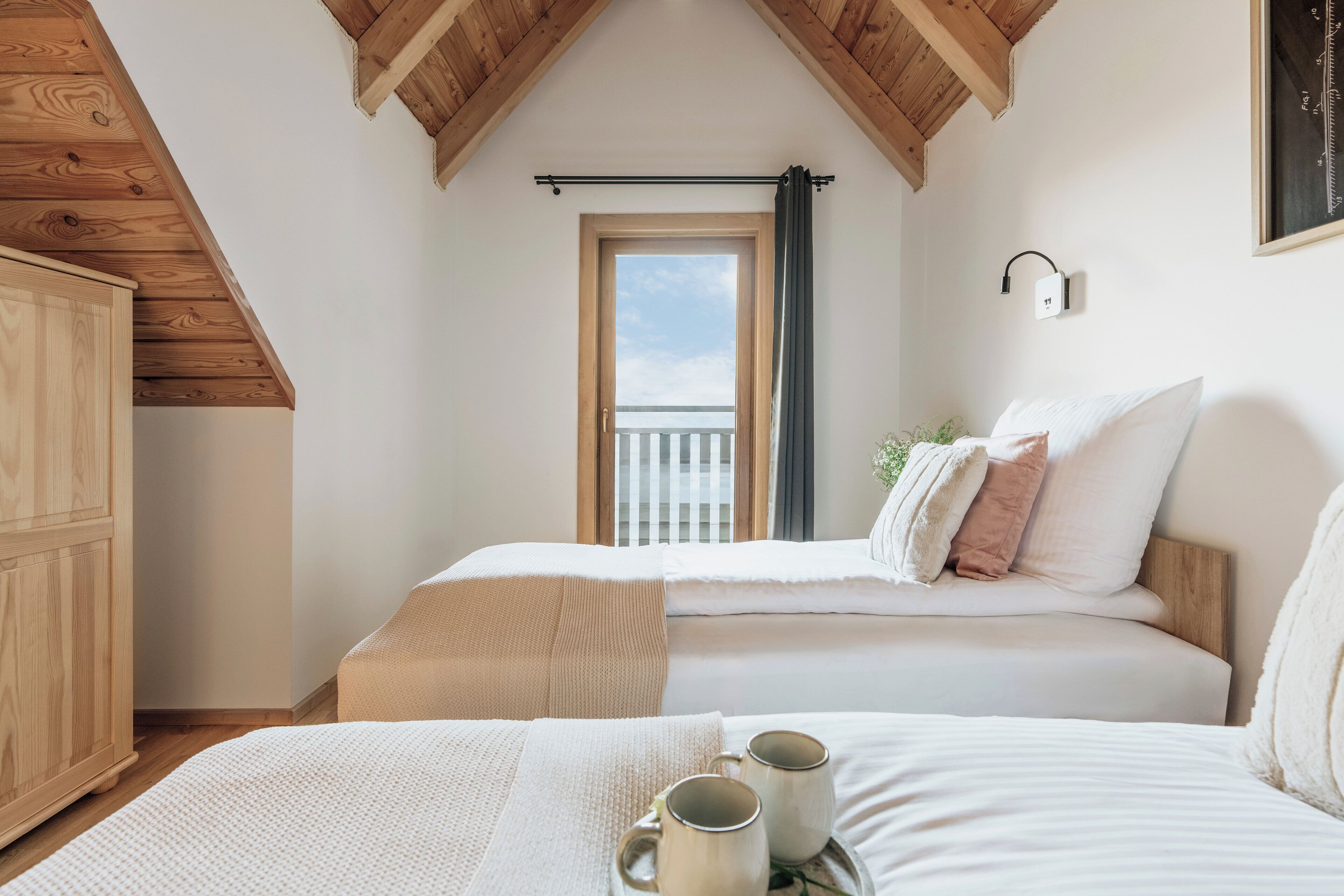 Une chambre avec un plafond en bois, un lit recouvert de draps blancs, une fenêtre donnant sur un beau paysage et une table de chevet sur laquelle reposent deux tasses.