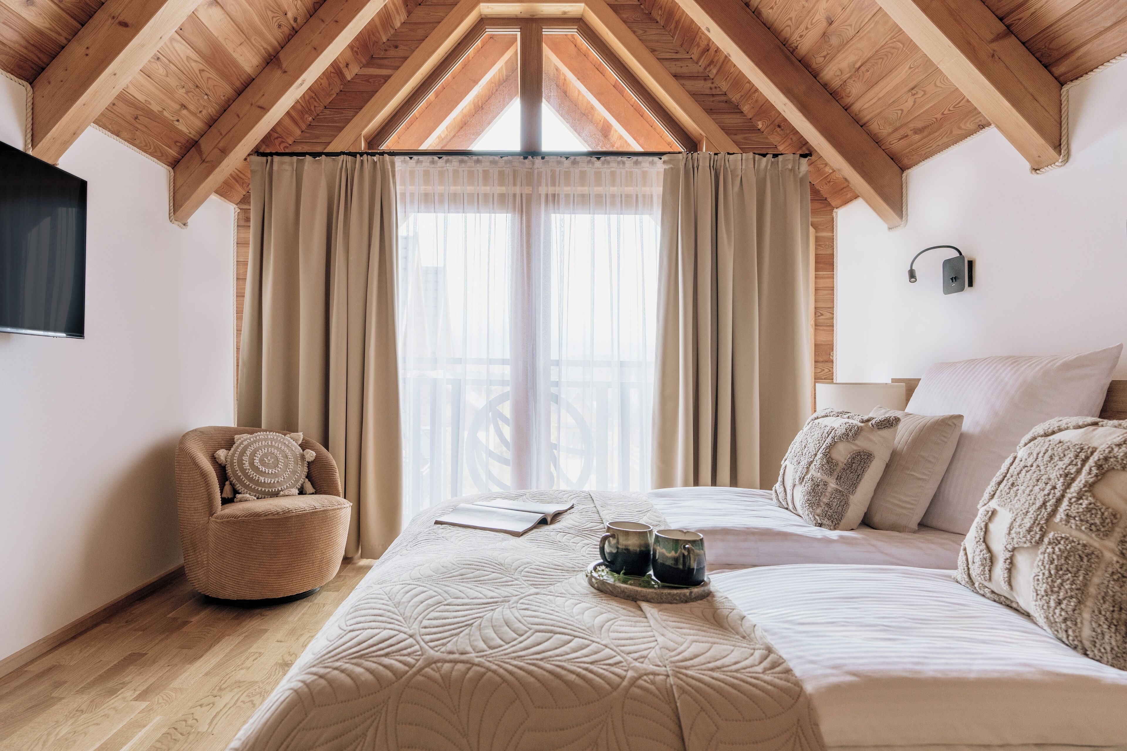 A bedroom with a large bed, a chair, a bedside table with two cups, and a window with curtains.