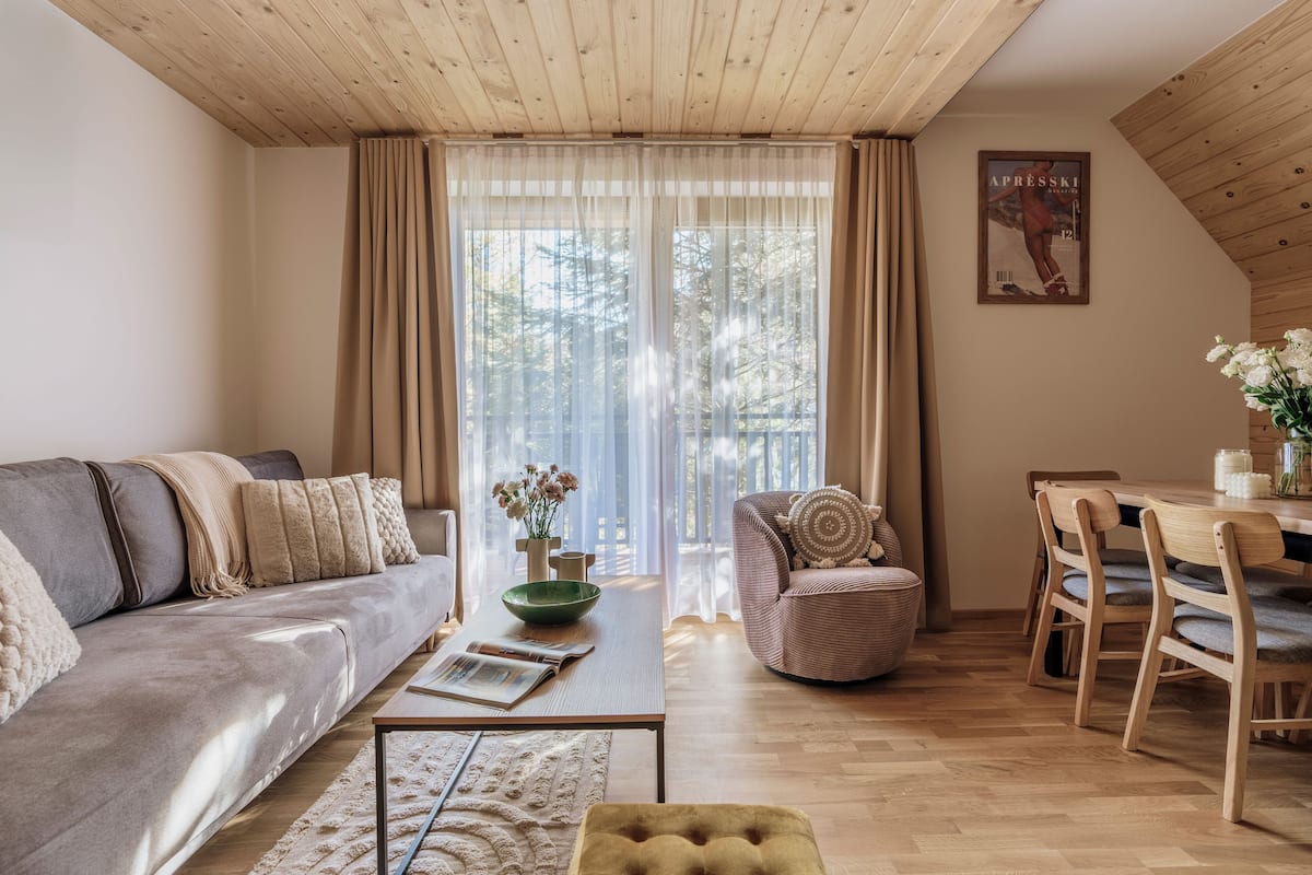 A modern living room with a sofa, a coffee table, a dining table with chairs, and a large window with curtains.