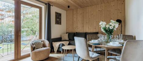 Ett modernt vardagsrum med ett träpanelat tak, en soffa, ett matbord för sex personer och utsikt över en trädgård genom stora fönster.