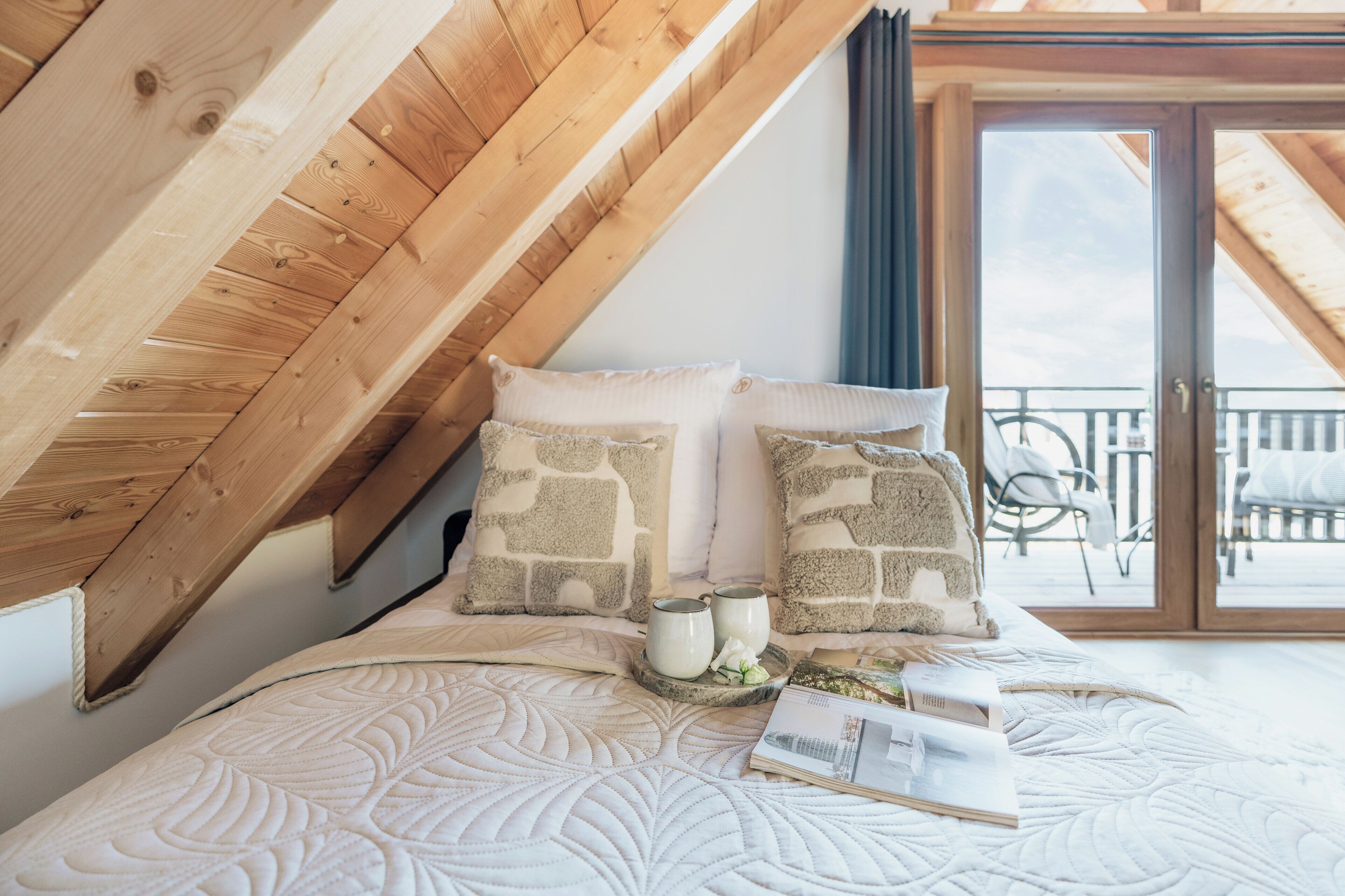 Een slaapkamer met schuine wanden, een bed met een gewatteerde spreidek, twee kussens, een nachtkastje met een boek en een mok, en uitzicht op het balkon.