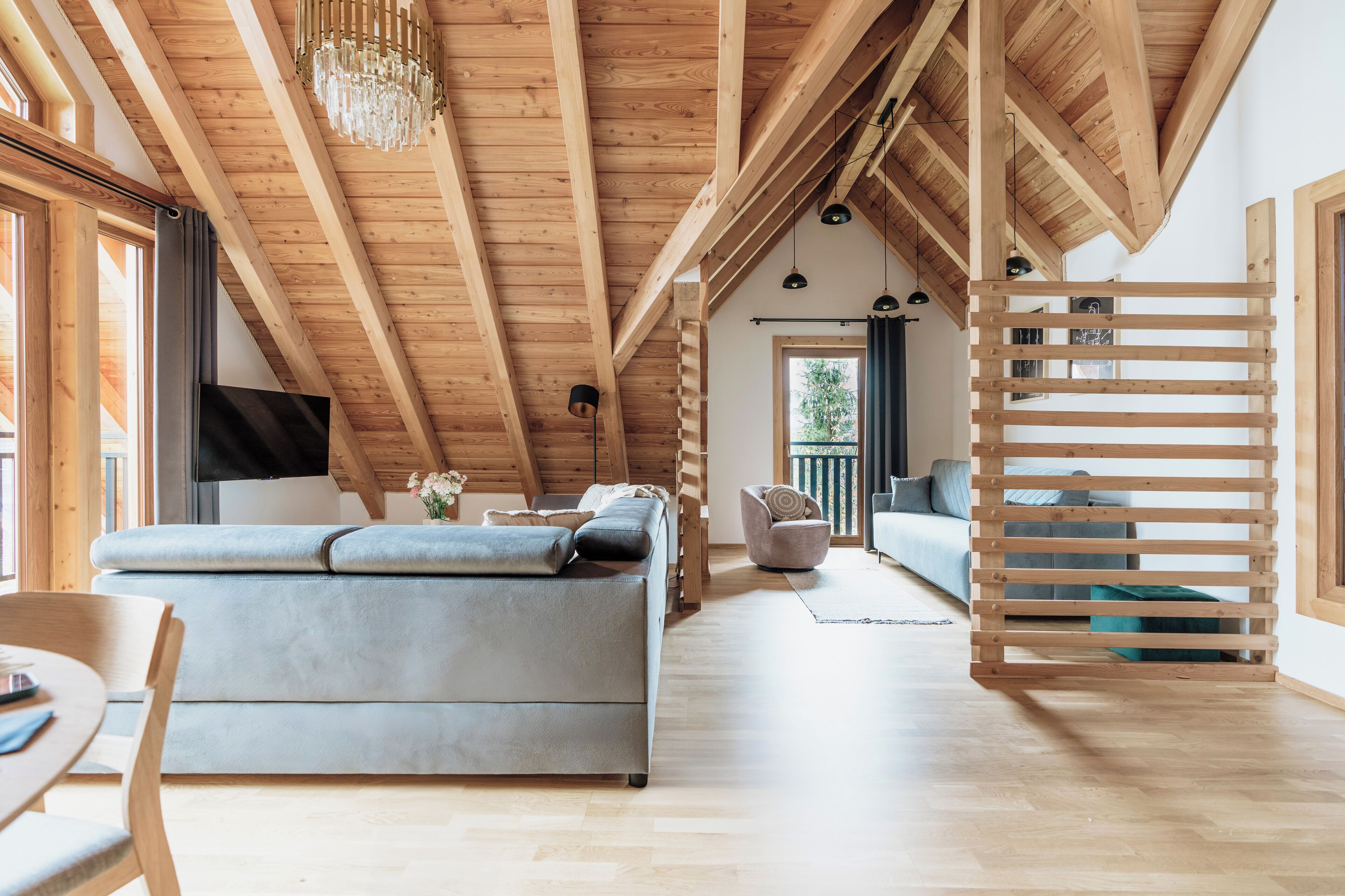 A spacious loft with exposed wooden beams, a grey sofa, a dining area, and a balcony with a wooden railing.
