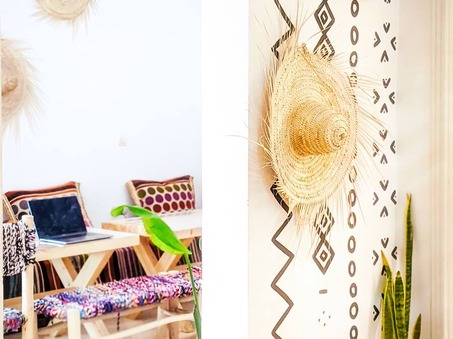 Una habitación con pared estampada, un tapiz tejido, una computadora portátil sobre una mesa de madera, una planta en maceta y una pequeña mesa con un cojín colorido.