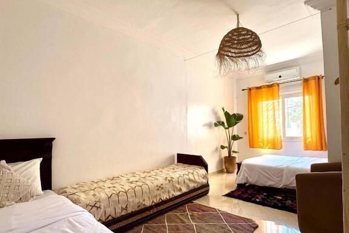 A hotel room with two beds, a large window with orange curtains, a hanging light fixture, and a potted plant.