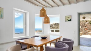 Un comedor con mesa y sillas de madera, ventanas amplias con vistas al océano, y lámparas colgantes de mimbre.