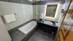 A modern bathroom with a glass-enclosed shower, a white bathtub, a black countertop with a sink, and a large mirror. - Rahaf Almashaer Hotel (Makkah)