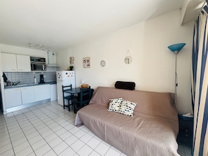 Ein kleines Schlafzimmer mit einem Bett, einem kleinen Tisch und einem Stuhl. Es gibt eine Küchenzeile mit einer Mikrowelle, einem Kühlschrank und einem Spülbecken.