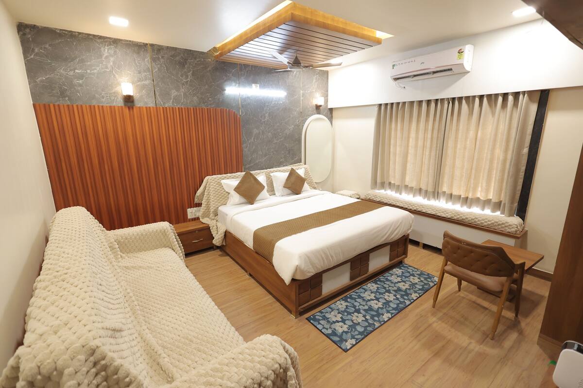 Une chambre d’hôtel moderne avec un grand lit, une tête de lit en bois, un coin salon avec un fauteuil, une petite table et une fenêtre avec des rideaux.