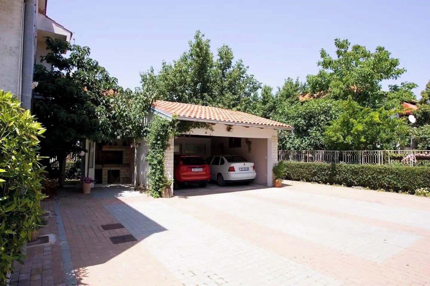 A covered parking area with two cars, surrounded by greenery and a white building.