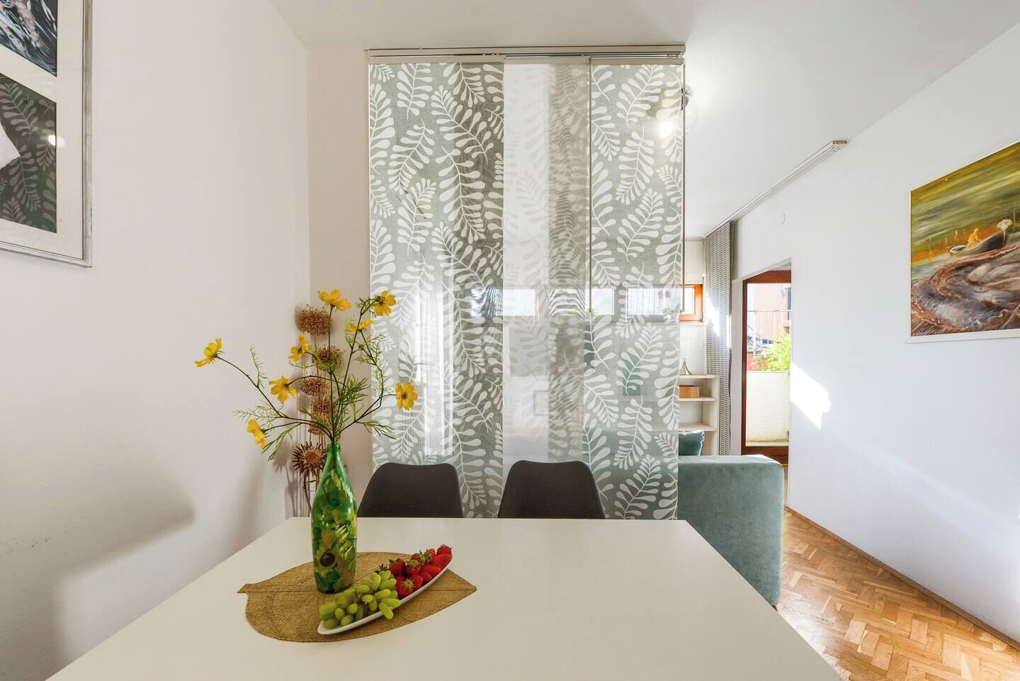 A dining area with a white table, a vase with yellow flowers, a fruit plate, and a patterned curtain.