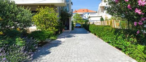 A paved pathway leading to a modern two-story building with balconies, surrounded by lush greenery and flowering plants.