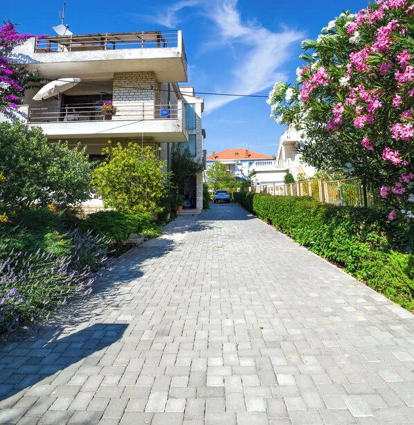 Un sendero pavimentado que conduce a un moderno edificio de dos pisos con balcones, rodeado de abundante vegetación y plantas en flor.