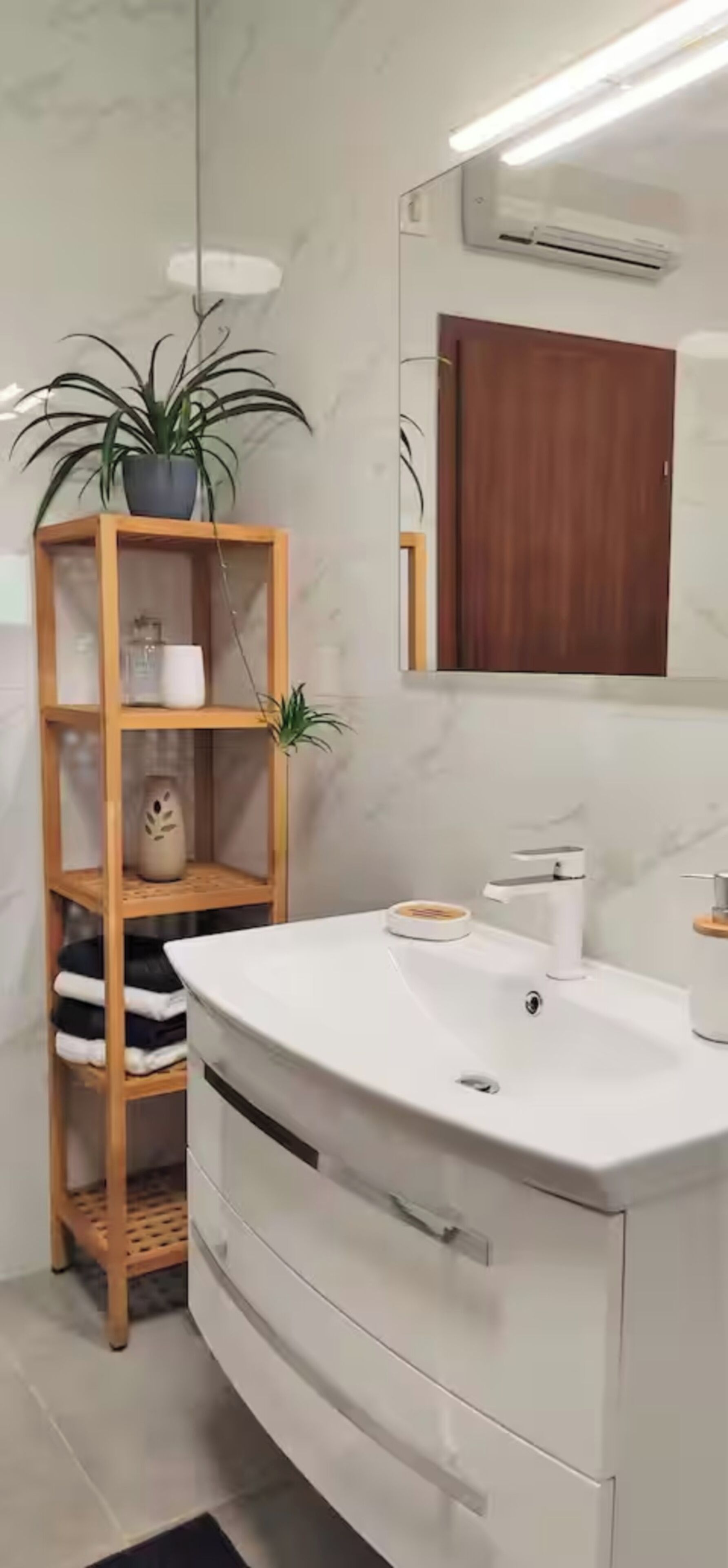 A modern bathroom with a white vanity, a wooden shelf with plants, and a mirror above the sink.